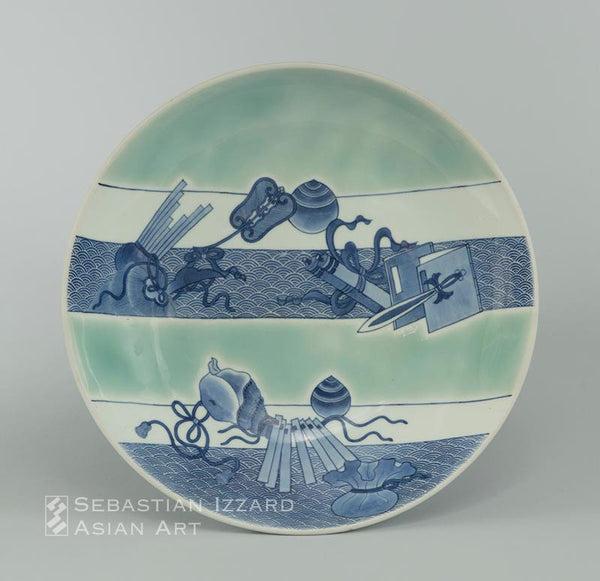 A SERVING DISH (SHAKUZARA) WITH THE ATTRIBUTES OF THE SEVEN GODS OF GOOD FORTUNE (TAKARAMONO)  Hizen ware, Nabeshima type: porcelain with underglaze blue and celadon Edo period, circa 1680 Diameter 32.8 cm